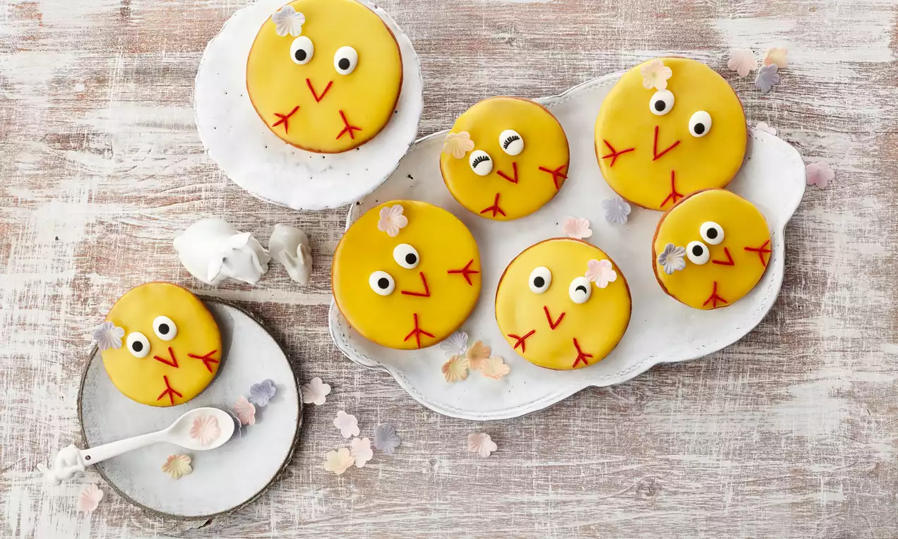 Oster-Amerikaner mit gelber Glasur, schwarzen und weißen Augen, rotem Schnabel und Zuckerblüten, arrangiert auf weißen Tellern