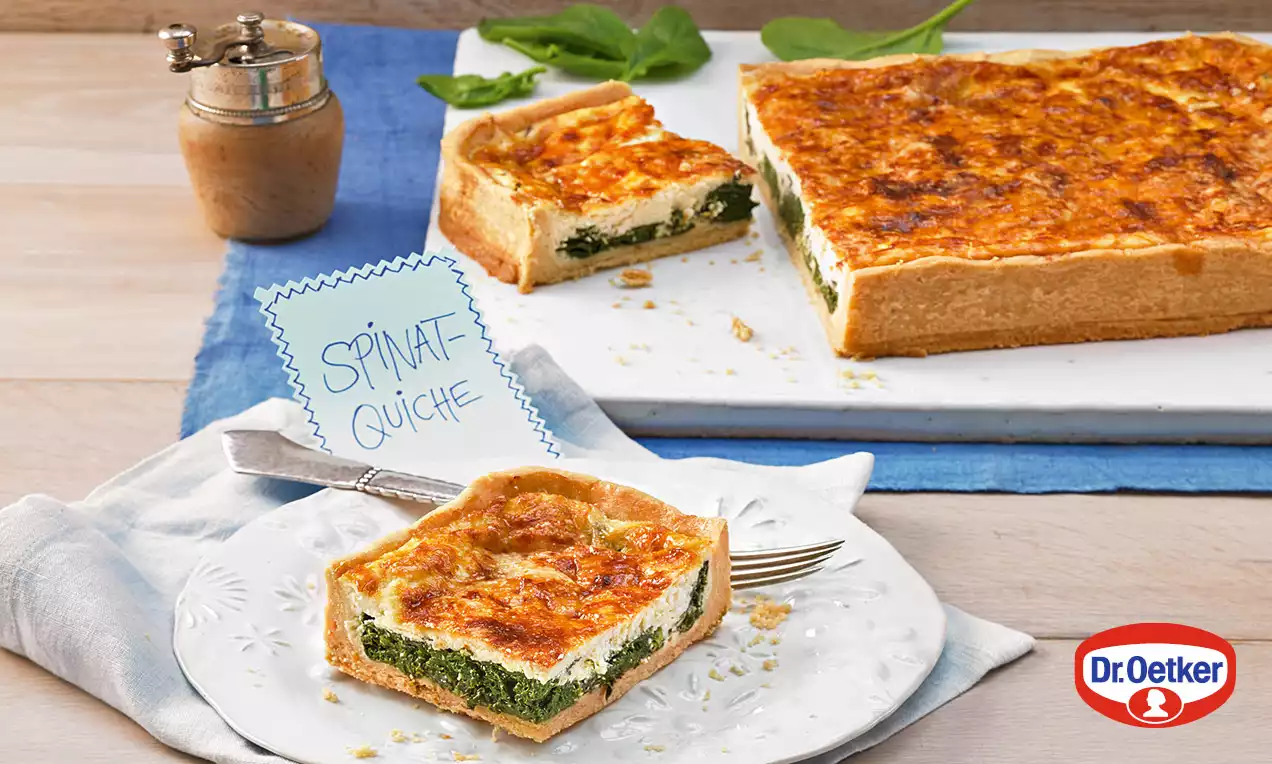 Špenátový quiche se zlatavou křupavou kůrkou, vrstvený krémovou náplní a špenátem, na bílém talíři vedle modrého ubrousku