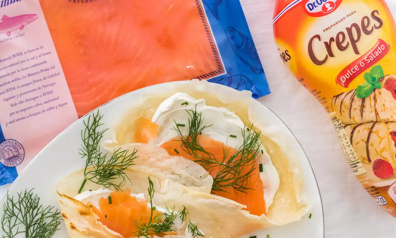 Crepes finas de tono dorado plegadas, con salmón ahumado naranja vibrante, crema blanca y eneldo fresco sobre un plato blanco
