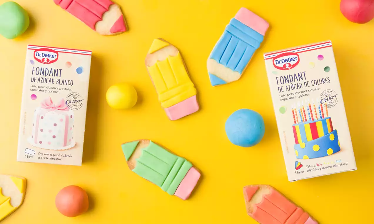 Galletas con forma de lápices de colores pastel, detalles en fondant y textura lisa distribuidas sobre fondo amarillo junto a cajas y esferas