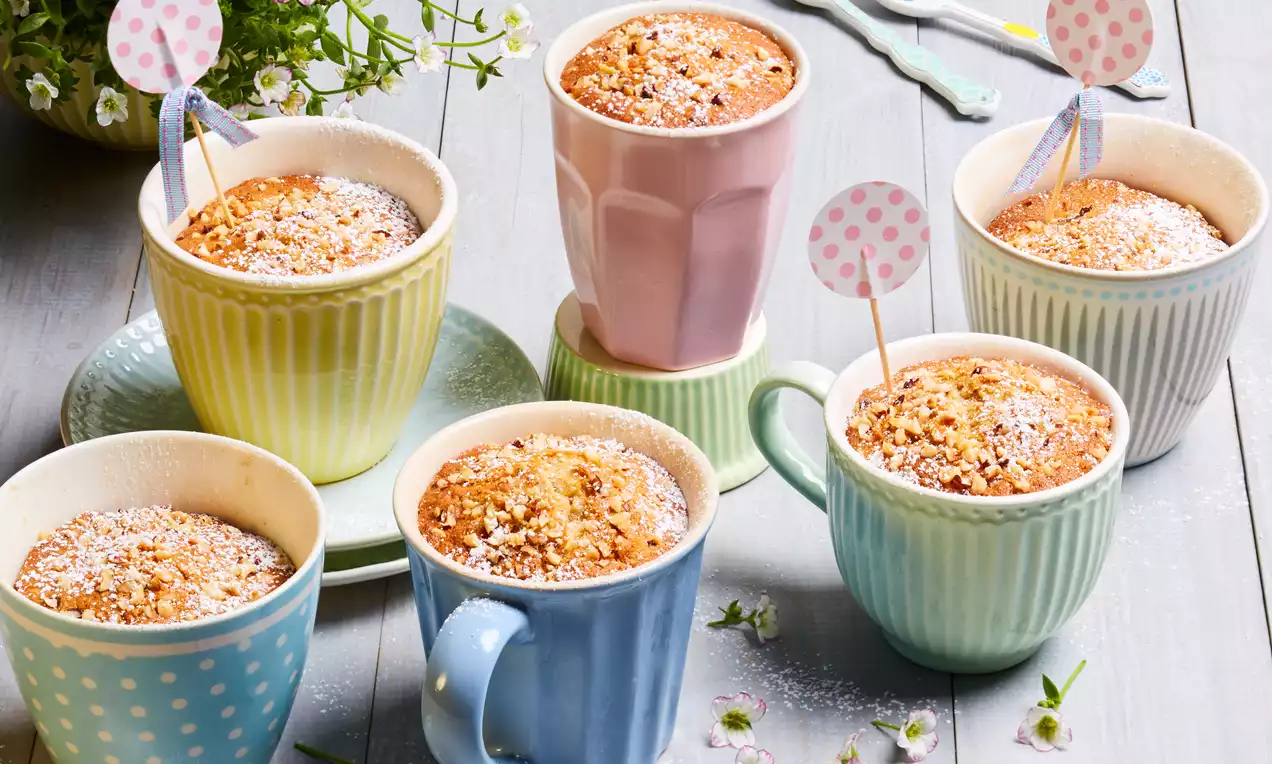 Goldbraune Bananen-Kokos-Tassenküchlein mit feiner Puderzuckerschicht in bunten Tassen, dekoriert mit Fähnchen und umgeben von Blüten
