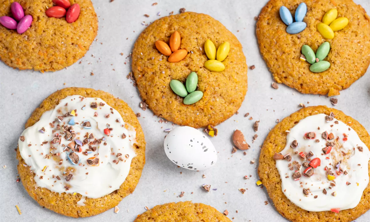 Mehki oranžno-rjavi piškoti z neenakomerno teksturo, okrašeni s pisanimi bonboni in belimi glazurami, razporejeni na papirju za peko