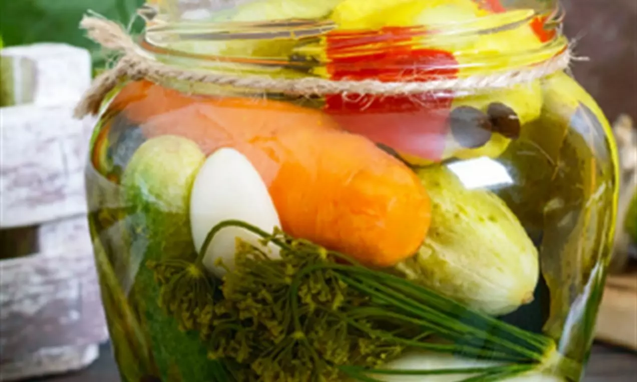 Разноцветни зеленчуци в стъклен буркан, потопени в златиста течност, с нежно зелени стръкчета копър и въже около гърлото на буркана