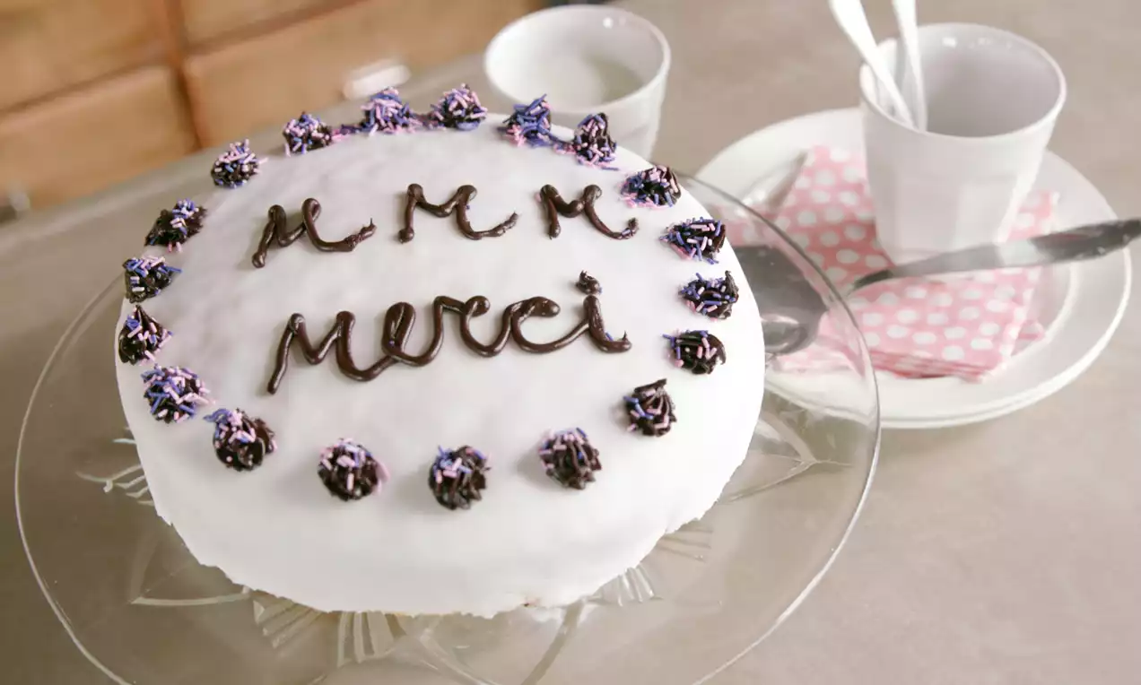 Ronde witte biscuitcake op een glazen taartplateau, met chocoladetext erop en paarse garnering omrand, naast kopjes en roze servetten