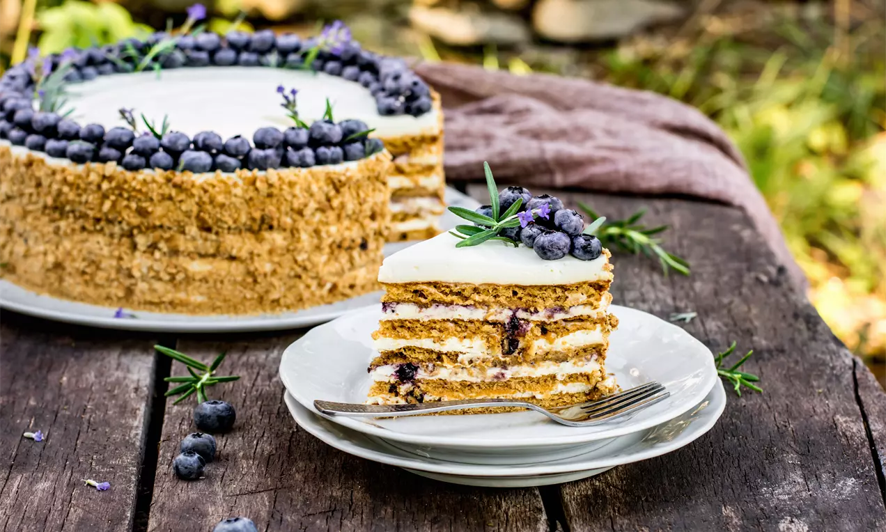 Медена торта с кремообразни слоеве бял чийзкейк пълнеж, украсена с пресни боровинки и стръкове розмарин върху тъмна дървена маса