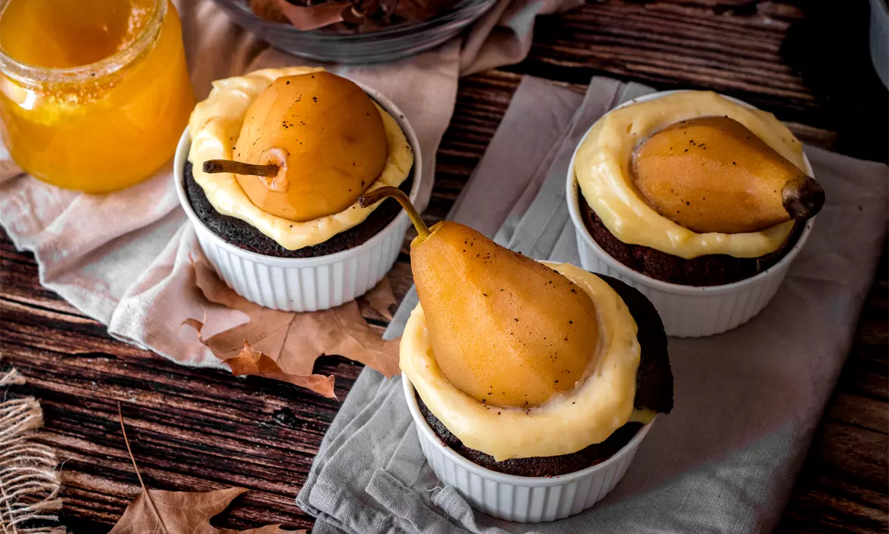 Шоколадово суфле с наситено кафяв цвят в бели купички, гарнирано с гладък ванилов крем и цяла поширана златисто-кафява круша върху текстил