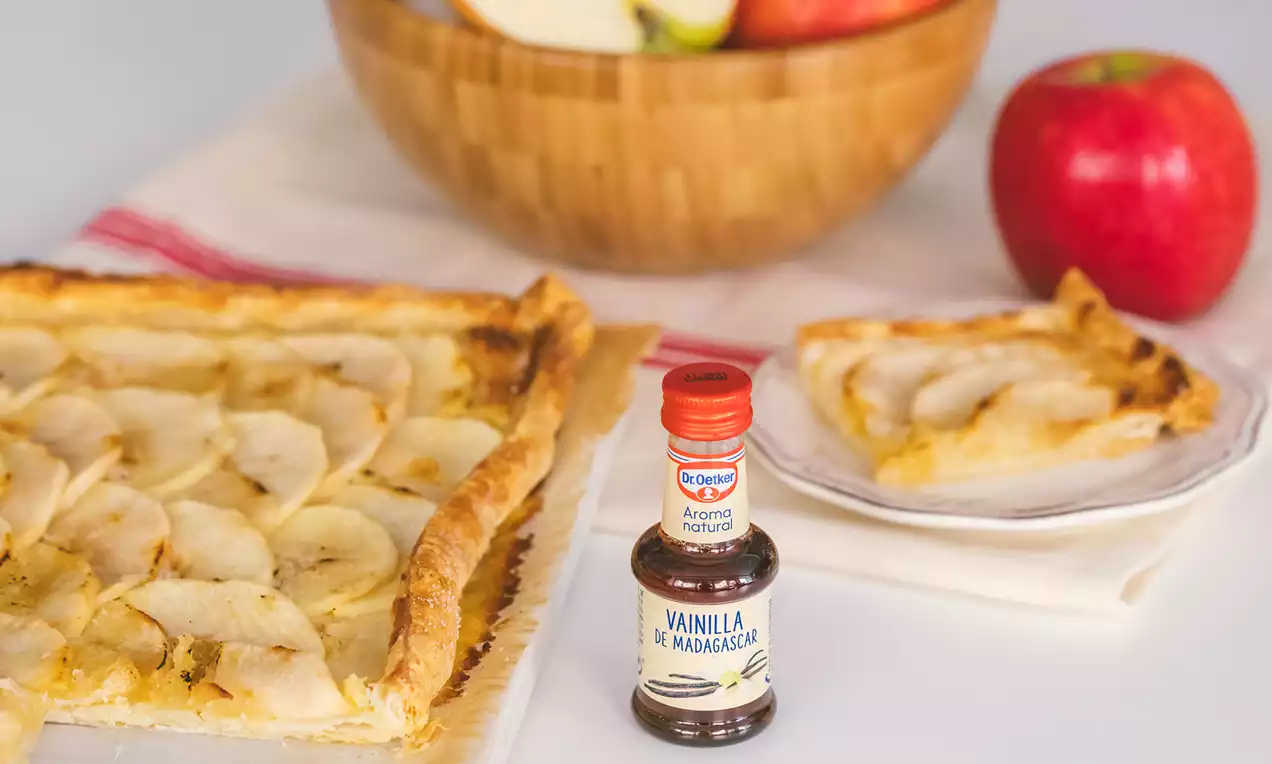 Tarta rectangular de hojaldre dorado con finas láminas de manzana dispuestas en filas, sobre mantel blanco junto a un cuenco de fruta y un frasco de vainilla
