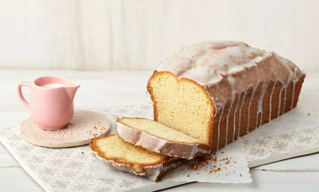 Веган сладкиш с правоъгълна форма, светлобежен отвътре, покрит с бяла глазура, поставен върху плоска чиния със светъл фон