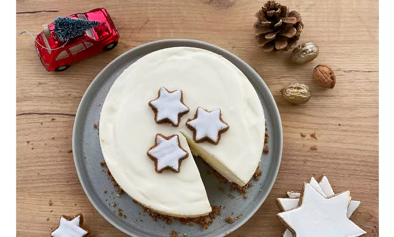 Runder Cheesecake mit glatter weißer Creme auf braunem Keksboden, dekoriert mit drei Zimtsternen, auf grauem Teller mit Weihnachtsdeko