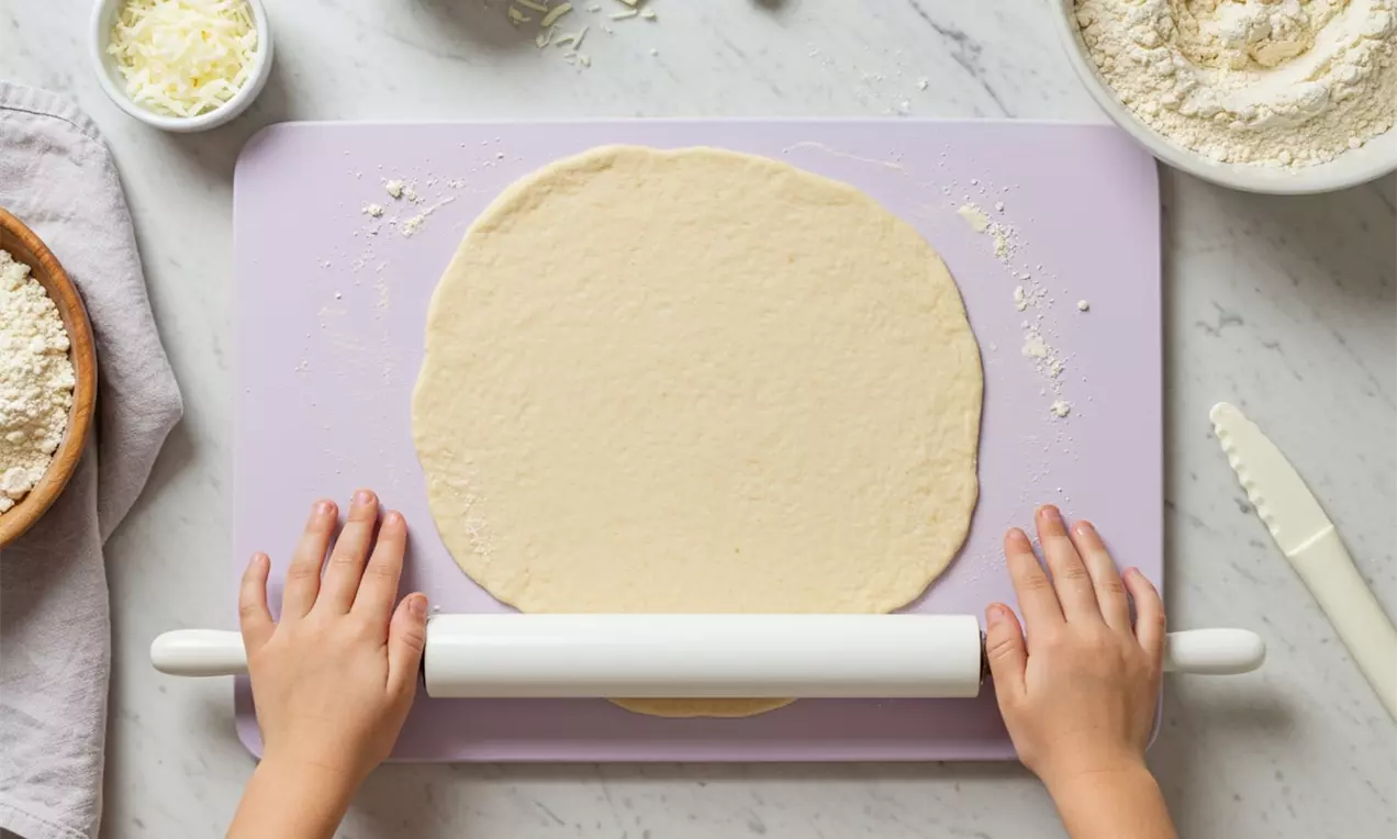 Children's hands rolling out smooth, pale beige pizza dough into a circular shape on a lavender mat surrounded by baking ingredients