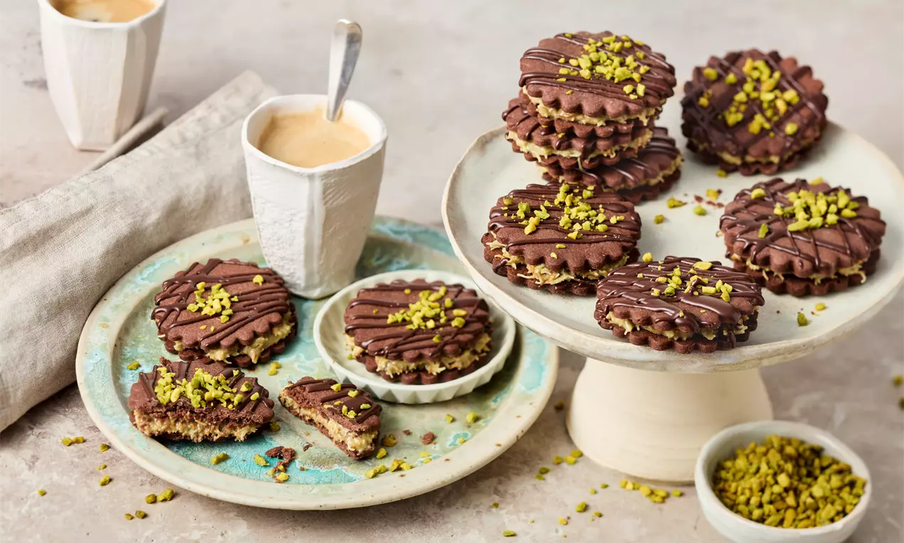 Chocolate cookies with ridged edges and drizzled chocolate, filled with rich cream and topped with vibrant crushed pistachios on plates