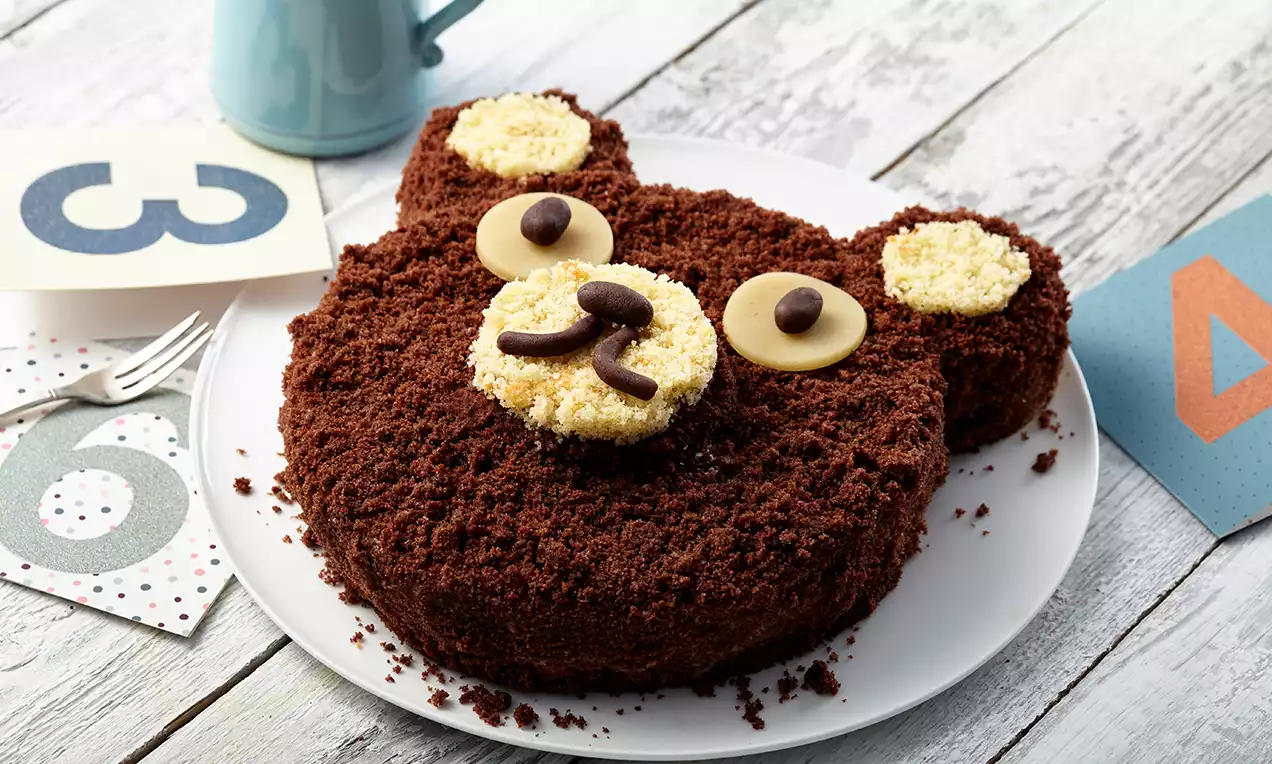 Runder Bärenkuchen aus braunen Kuchenkrümeln mit cremefarbenen Ohren, Schokoladenaugen und Nase auf weißem Teller, dekoriert mit Zahlenkarten