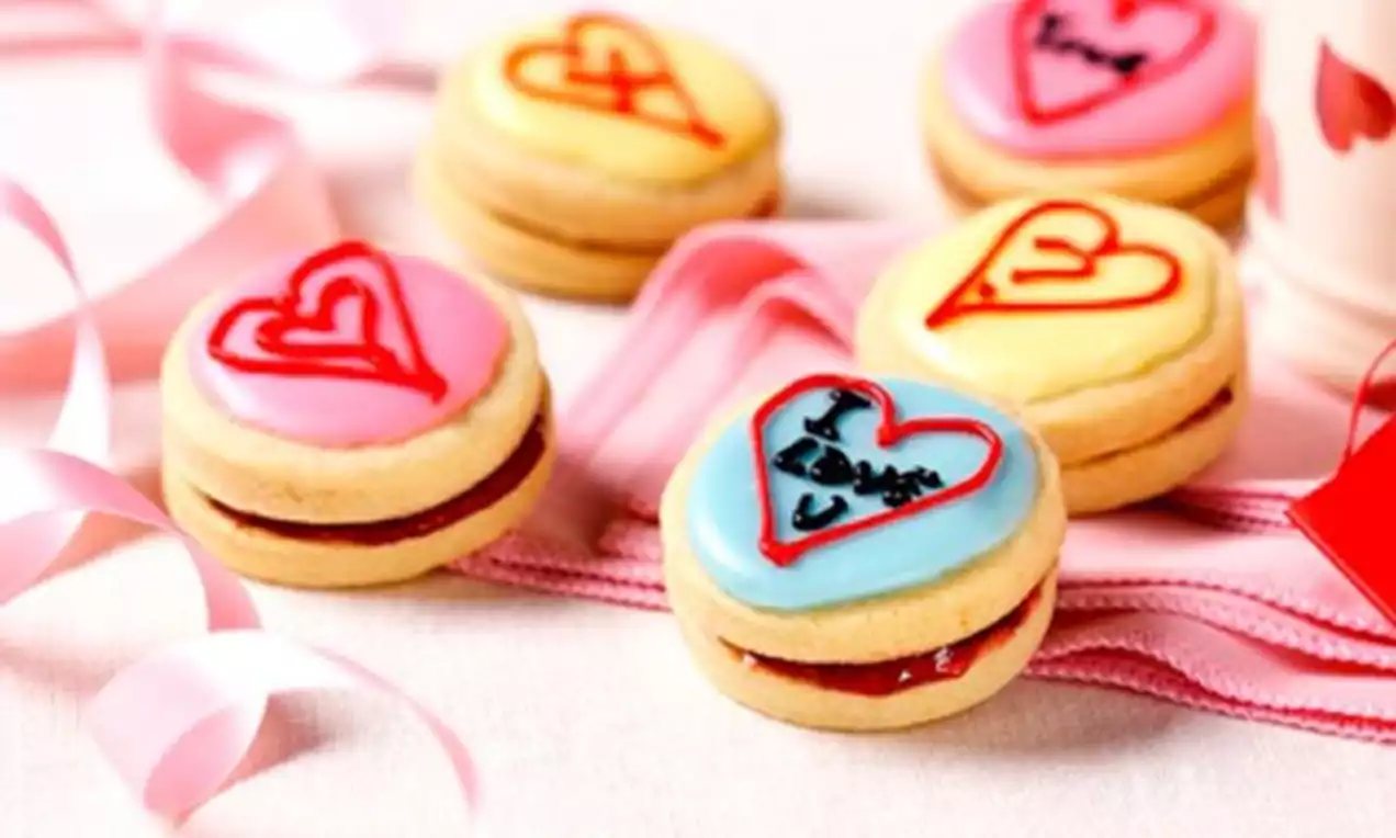 Lyse småkaker med et lag rød syltetøy, dekorert med fargede hjerter på glasur ligger på rosa bånd mot en lys bakgrunn