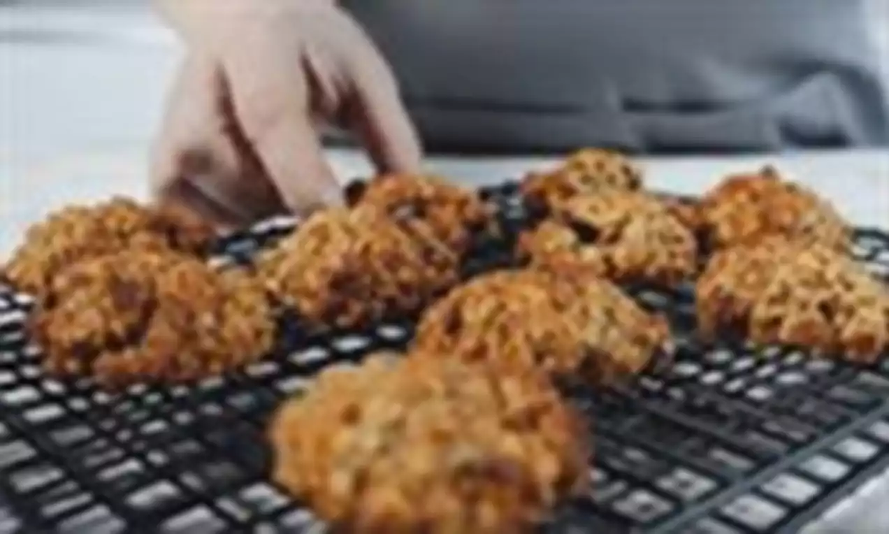 Zeltaini rūsgani, nevienmērīgas formas cepumi ar kraukšķīgu tekstūru izkārtoti uz melna metāla režģa, fonā redzama roka