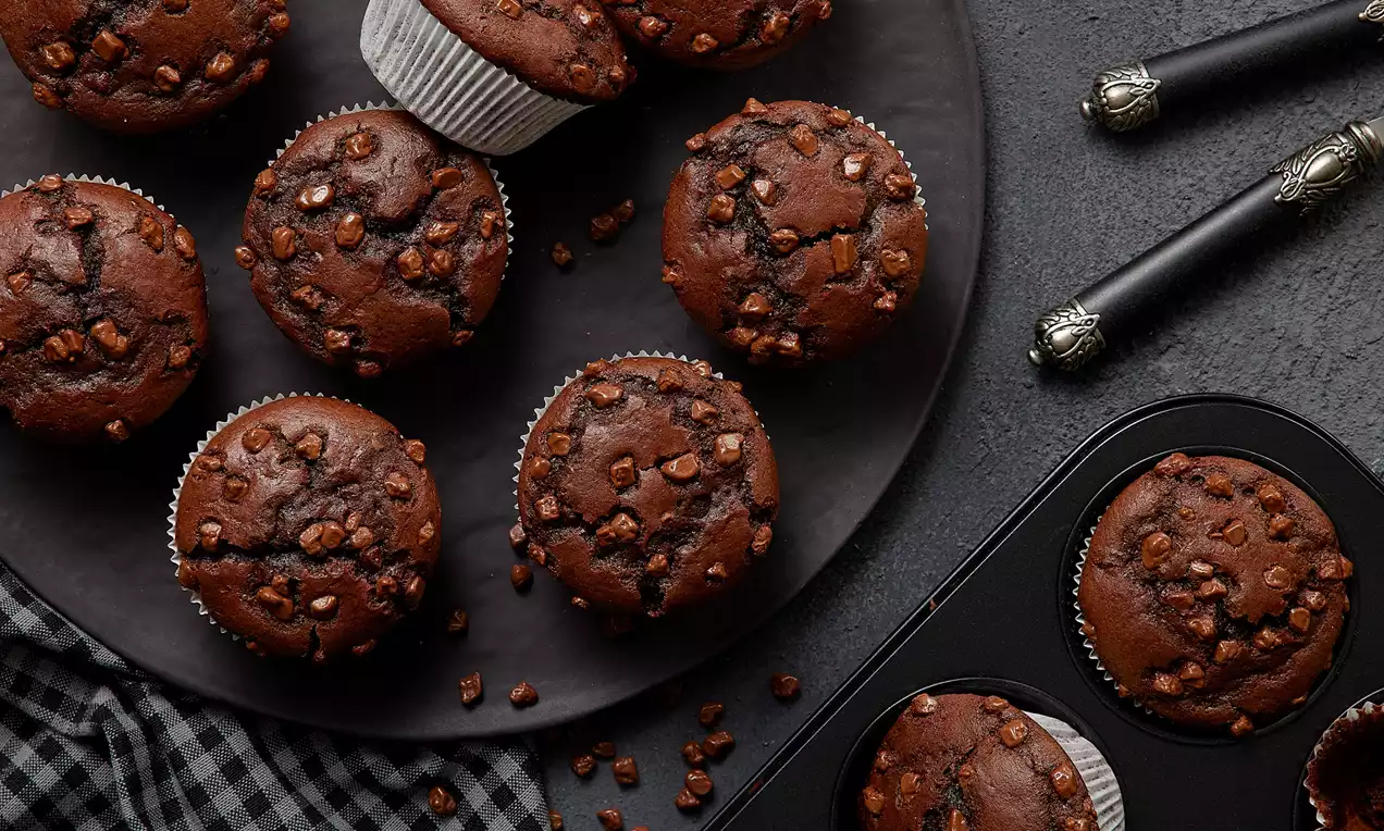 Koyu kahverengi çıtır çikolata parçalarıyla süslenmiş muffinler yuvarlak bir servis tabağında ve zarif siyah detaylarla birlikte sunulmuş