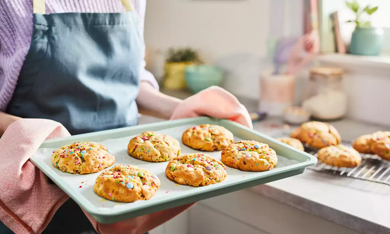 Warm, thick cookies soft and golden with a crinkled surface, studded with multicoloured candy pieces on a mint green baking tray