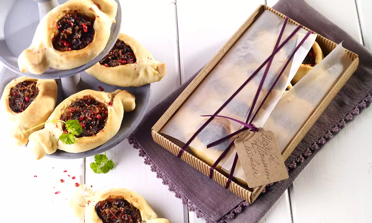 Aranyszínű, ívelt tésztabatyuk közepén lilás szilvás-zöldfűszeres töltelékkel, fehér porcelánon és lila szövettel díszítve