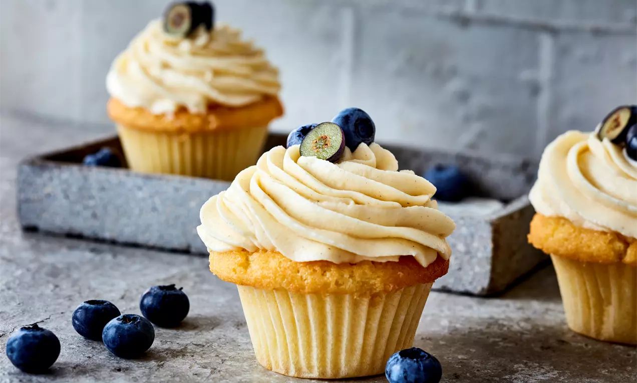 Кремообразна ванилова глазура със светъл оттенък, оформена на вълни върху меко златисто мъфиново тесто, гарнирана с сини боровинки