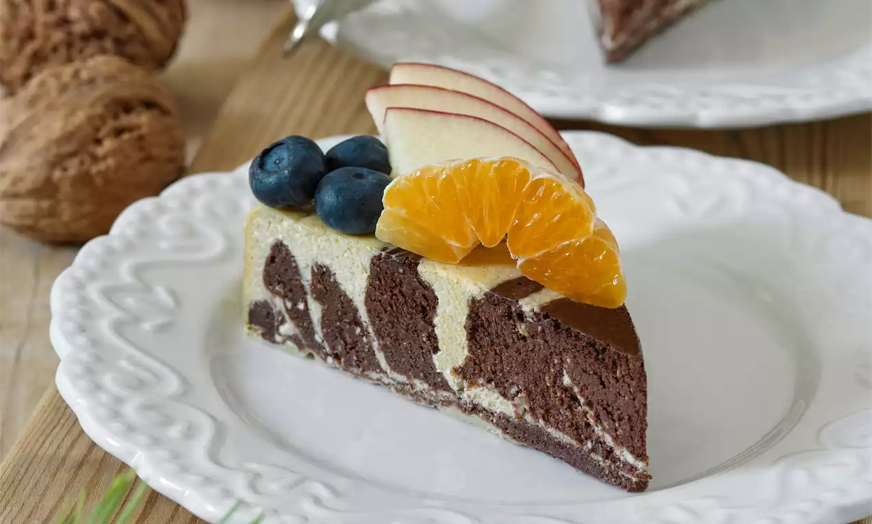 Pudingový koláč s mramorovým vzorom čokolády a vanilky, ozdobený kúskami mandarínky, plátkami jablka a čučoriedkami na bielom tanieri