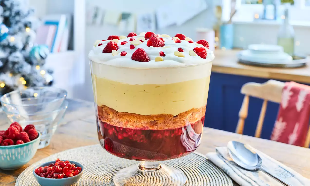 Layered vegan trifle in a glass bowl with red jelly base, golden sponge, pale custard, white cream topping and raspberries dotted on top