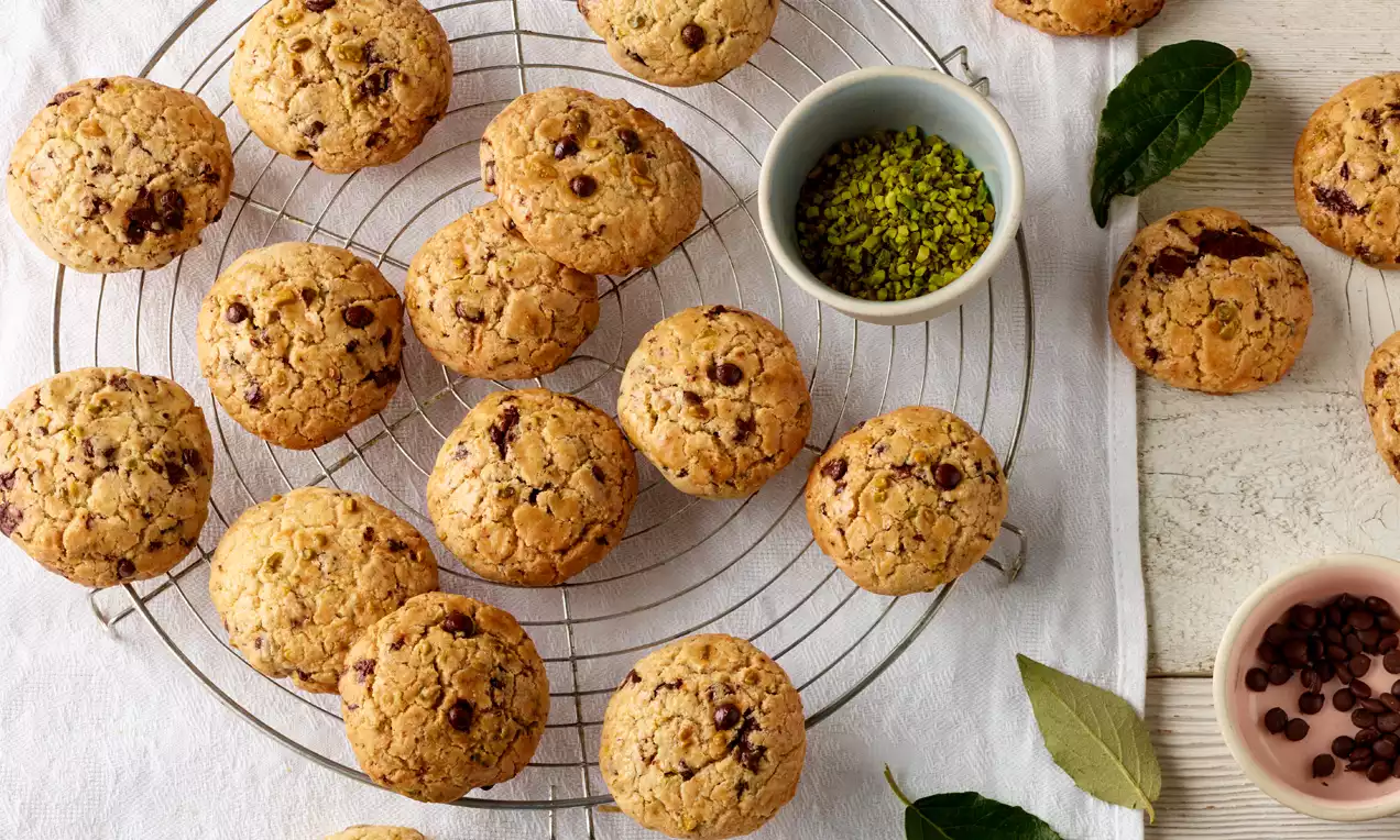 Runde Cookies mit goldbrauner Oberfläche, dunkler Schokolade und grünen Pistazienstücken auf einem Gitter, umgeben von Schalen und Blättern
