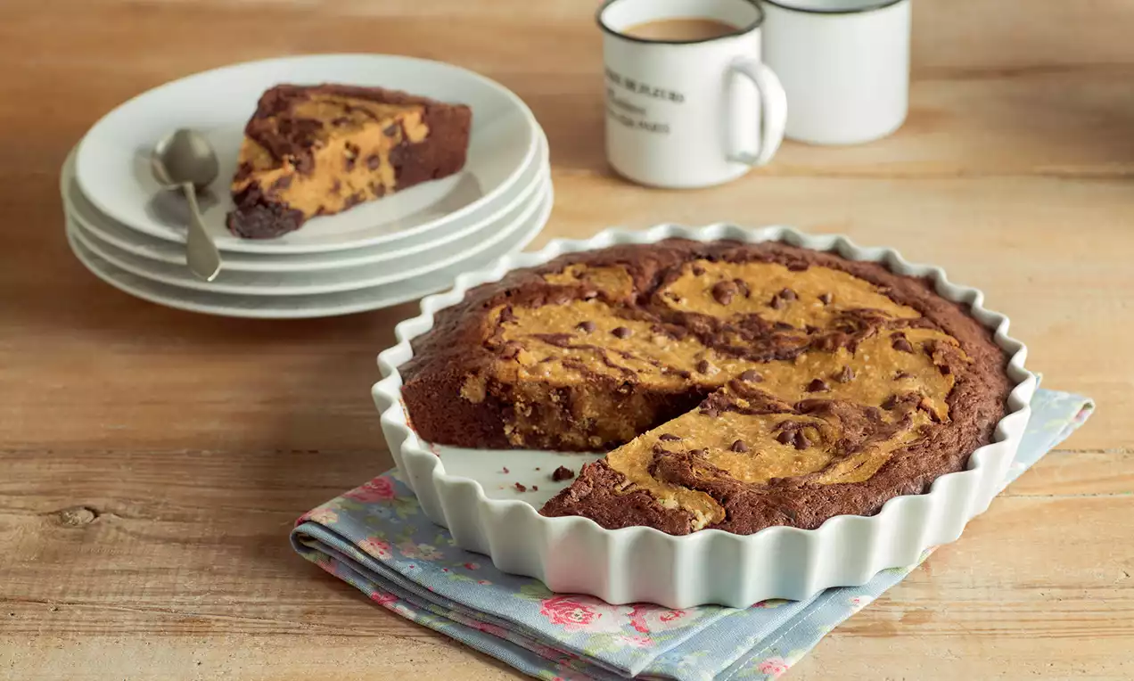 Tarta de chocolate oscuro y masa dorada con chips, en molde rizado blanco sobre paño estampado, junto a platos y tazas de cerámica