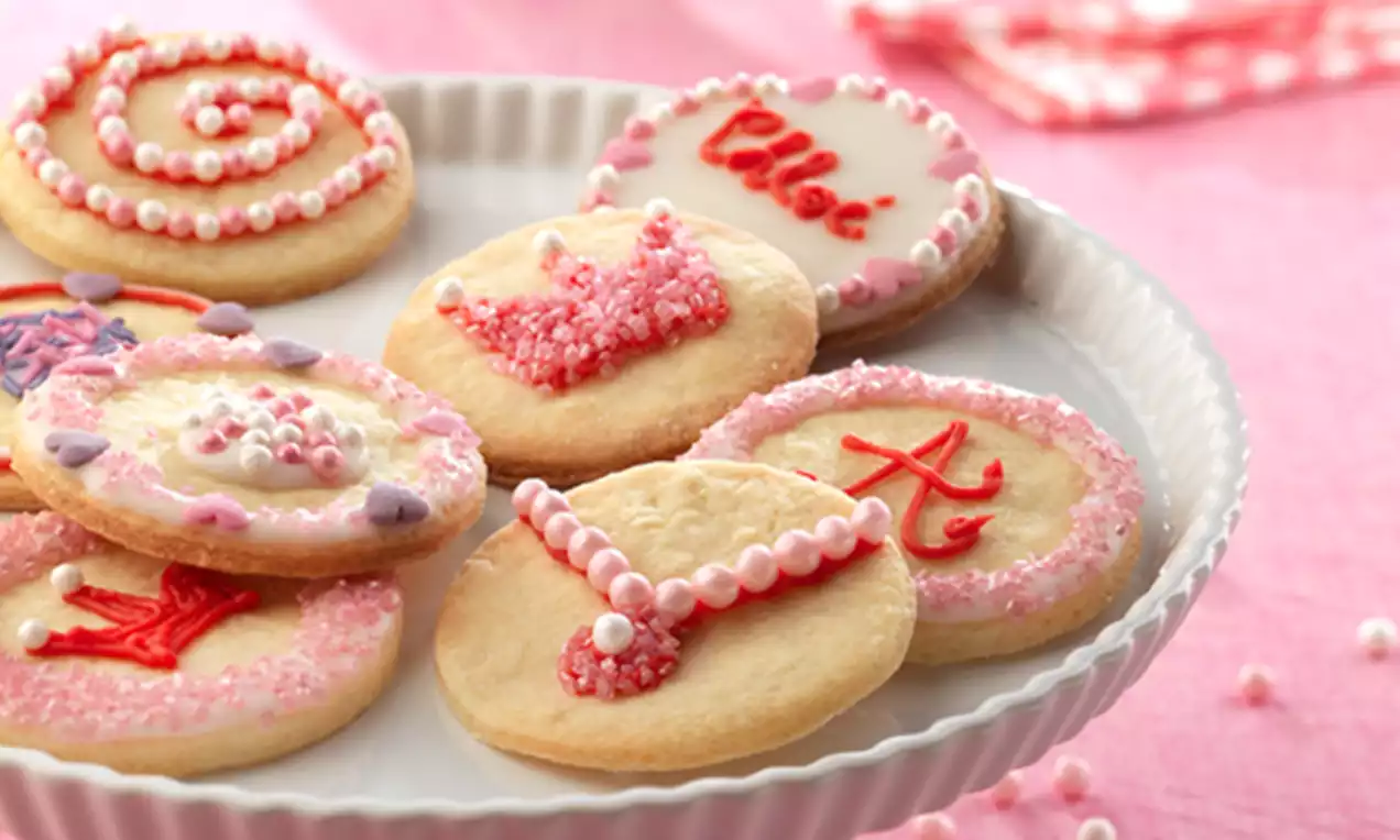 Galletas redondas doradas decoradas con glasas rosas y blancas, pequeños detalles perlados y coronas sobre un plato blanco ribeteado