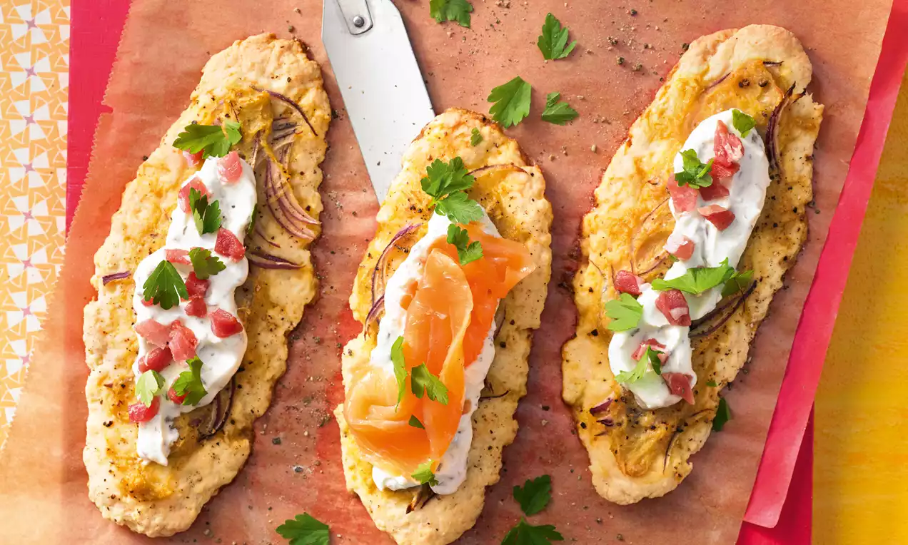 Goldbraune Kartoffelfladen in länglicher Form mit Sauerrahm, roten Zwiebeln, Lachs, Kräutern und Speckstücken auf rotem Backpapier