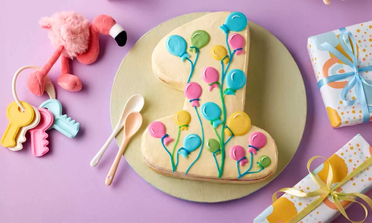 Gebäck in Form einer gelben Eins, verziert mit bunten Luftballon-Motiven, auf hellgrüner Platte, dekoriert mit Spielzeug und Geschenken