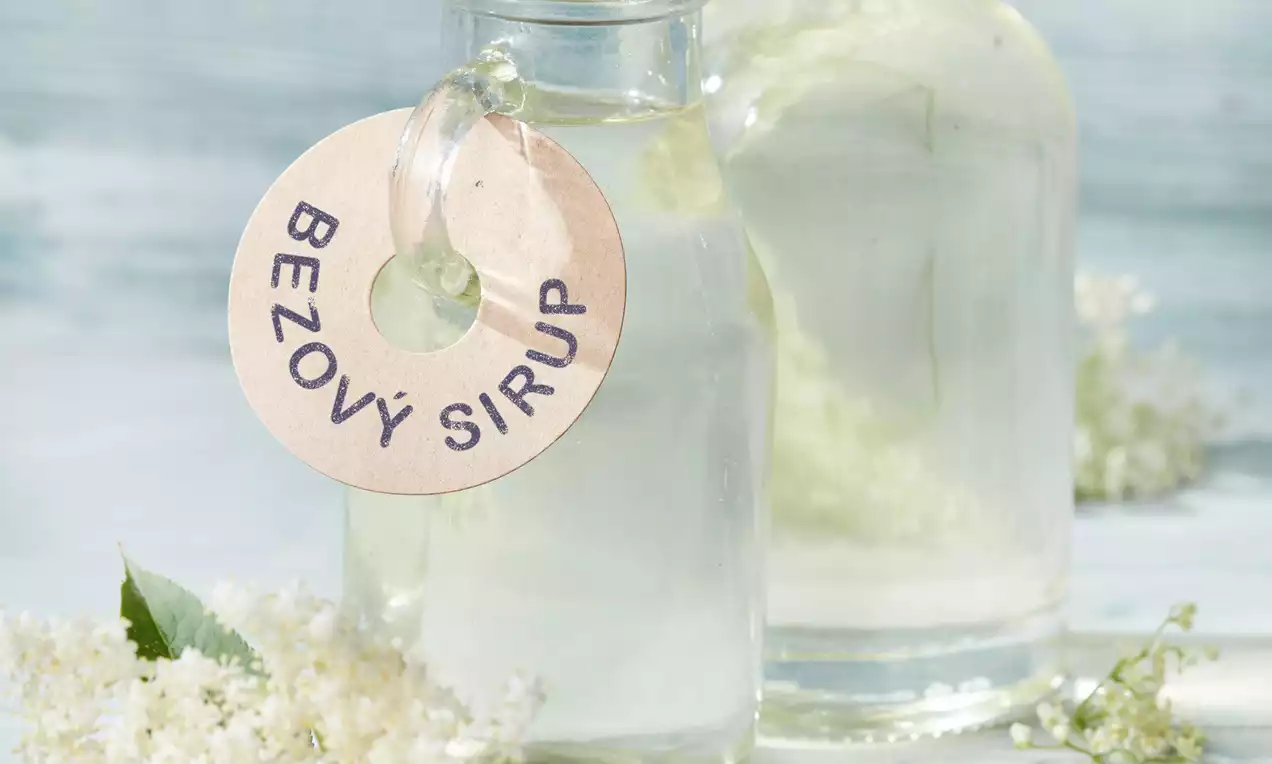 Průhledný bezový sirup ve skleněné lahvi s kovovým víčkem, ozdobený štítkem, obklopený drobnými bílými květy na světle modrém pozadí