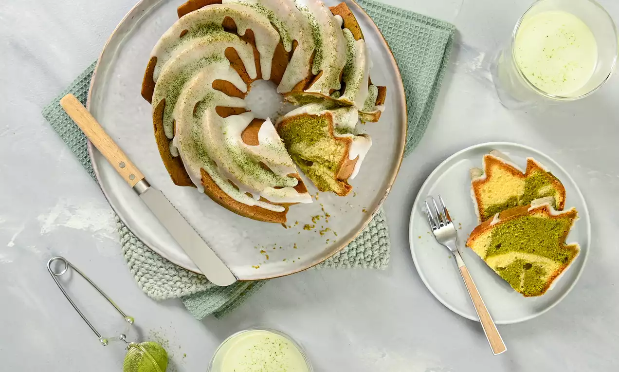 Plant-based matcha Bundt cake with swirls of green and golden layers, drizzled with white icing on a white plate with a knife and matching slices nearby