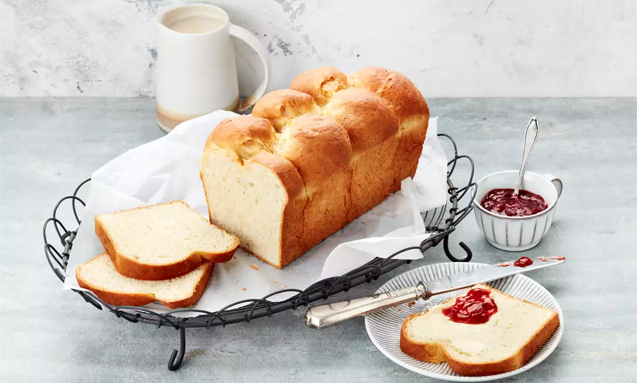 Topao, zlatnosmeđi kruh brioš s mekanom, glatkom teksturom na metalnom pladnju, uz kriške raspoređene i poslužene s marmeladom