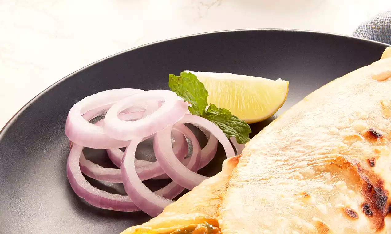 Chicken Kathi Roll with golden toasted flatbread wrapped around spiced chicken filling, served on a black plate with onion rings, lime wedge and a bowl of red cherry tomatoes in the background