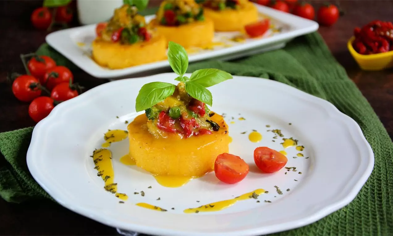 Na belom tanjiru nalazi se zlatnožuti medaljon od palente, ukrašen svežim povrćem, listićem bosiljka i dekorativnim kapljicama sosa