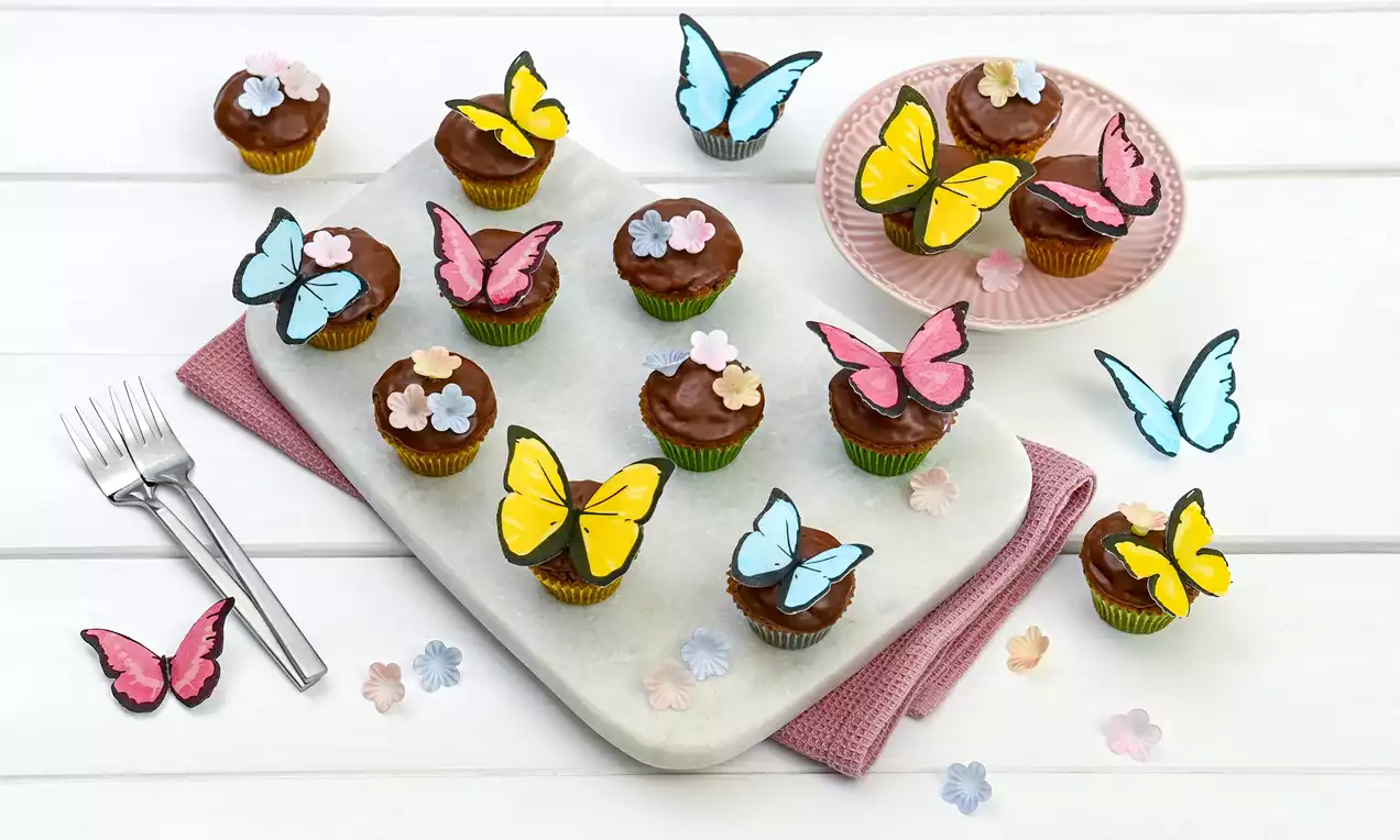 Mini-Schoko-Muffins mit glänzender Schokoladenglasur, bunten Zuckerblumen und Dekoschmetterlingen auf Tablett und rosa Teller angerichtet