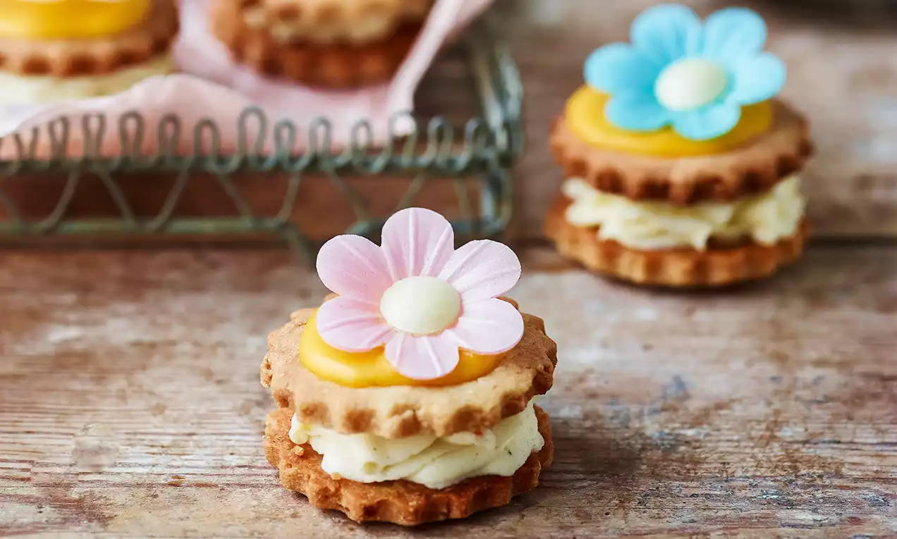 Små medaljer av lysebrun kjeks med kremfyll og gul sitronpassionsskum, toppet med rosa og blå blomster på rustikk bordflate