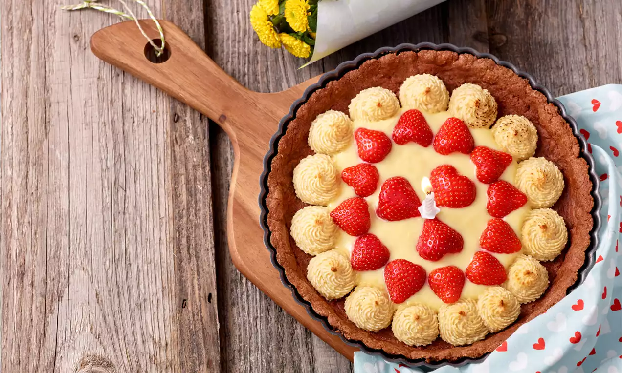 Erdbeer-Tarte mit goldbraunem Mürbeteigboden, gelber Cremefüllung, roten Erdbeerstücken und hellen Marzipanrosen auf einem Holzbrett