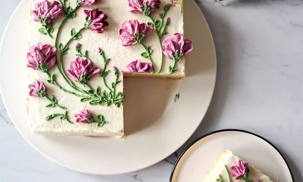 Квадратен кейк с бяла маслена глазура, декориран с розови цветя и зелени листа от крем, поставен върху светла чиния на мраморен фон
