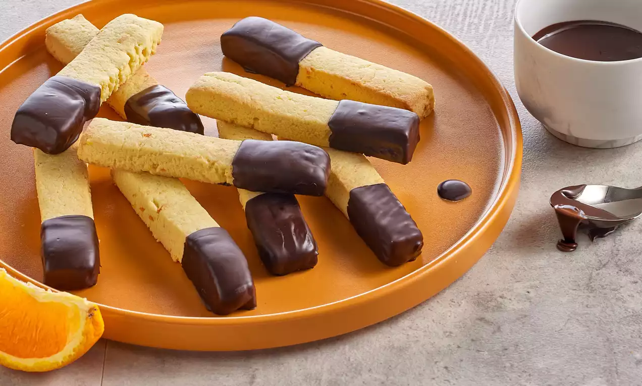 Portakal dilimi eşliğinde turuncu bir tabakta sunulan uçları çikolataya batırılmış altın sarısı çıtır çubuk kurabiyeler