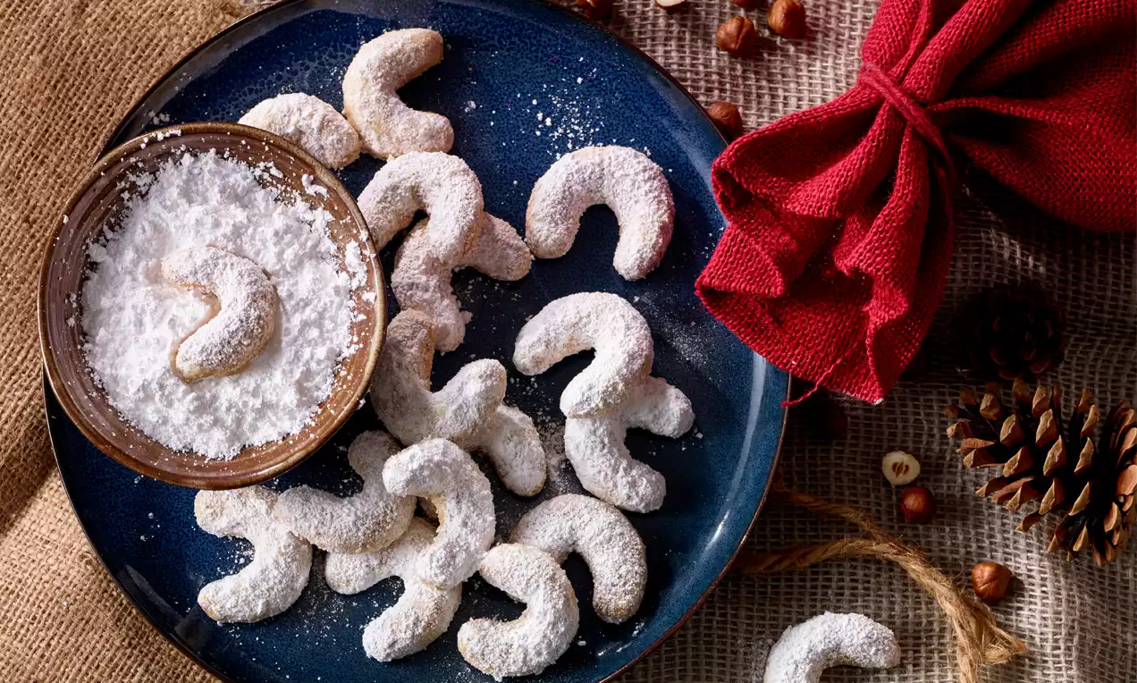 Goldbraune Vanillekipferl mit zarter Puderzuckerschicht, geschwungen angeordnet auf blauem Teller neben Holzschale mit Puderzucker