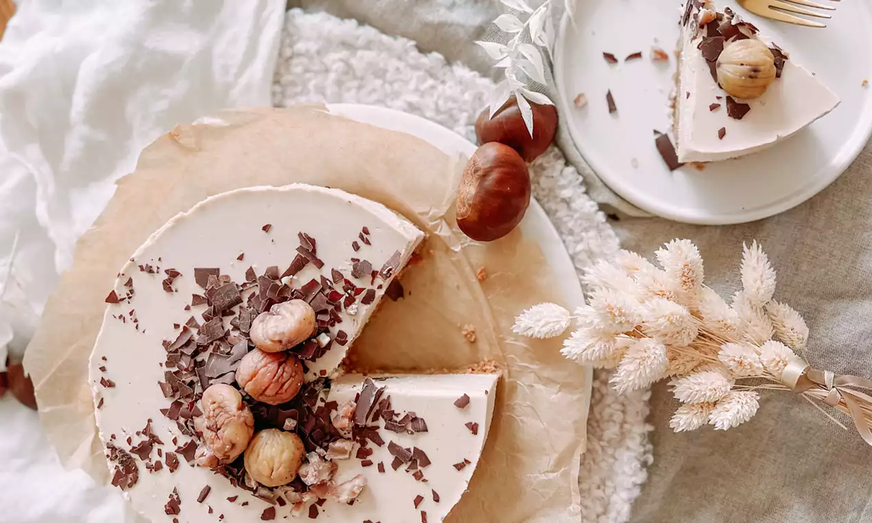 Marroni Cheesecake mit heller, cremiger Oberfläche, dekoriert mit ganzen Kastanien und dunklen Schokosplittern auf einem Teller mit Trockenblumen