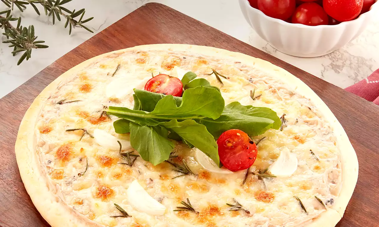 A golden round Pizza Bianca with a creamy surface topped with fresh arugula, red cherry tomatoes, and herbs on a wooden board