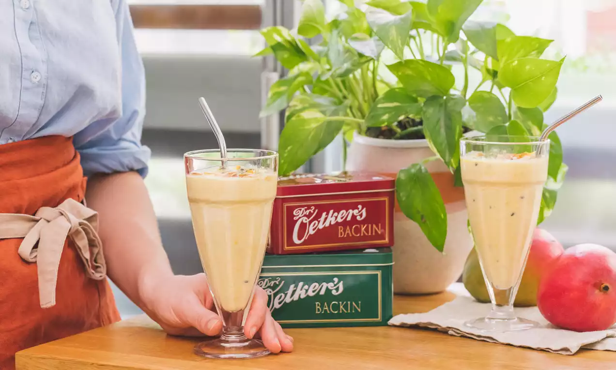 Batido amarillo cremoso en vaso alto con pajita metálica, junto a cajas de colores, frutas frescas y una planta verde sobre madera clara