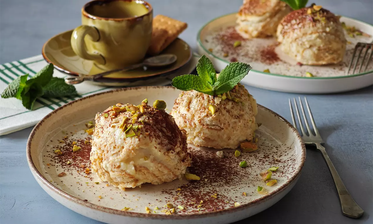 Две кремообразни топки ванилова бисквитена торта с поръска какао и натрошени шамфъстъци върху керамична чиния, украсени с свежи листа мента