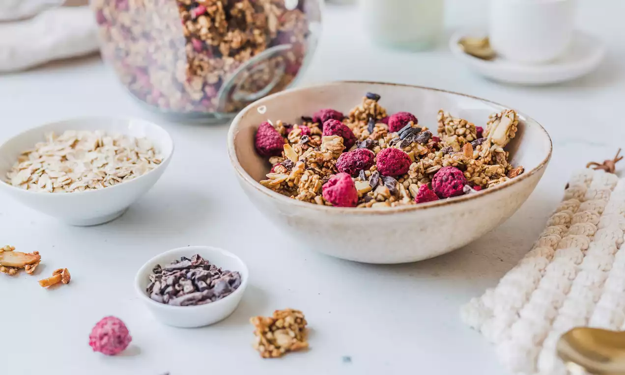 Knuspriges Granola mit goldbraunen Stücken und leuchtend roten Himbeeren in einer beige-grauen Schale, daneben Zutaten in kleinen Schälchen