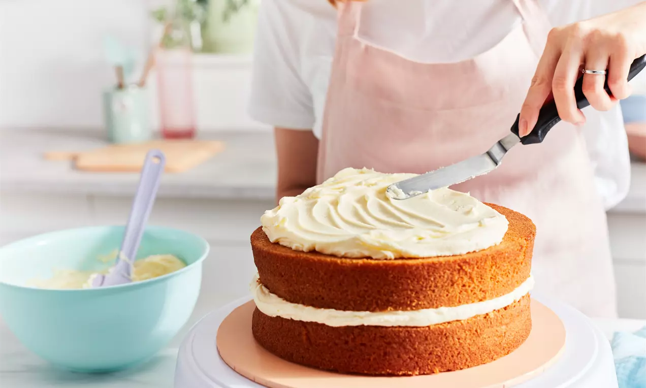 Висока двуслойна торта с равномерни златисто-кафяви пластове, покрита със светъл крем с мека текстура, оформян с шпатула