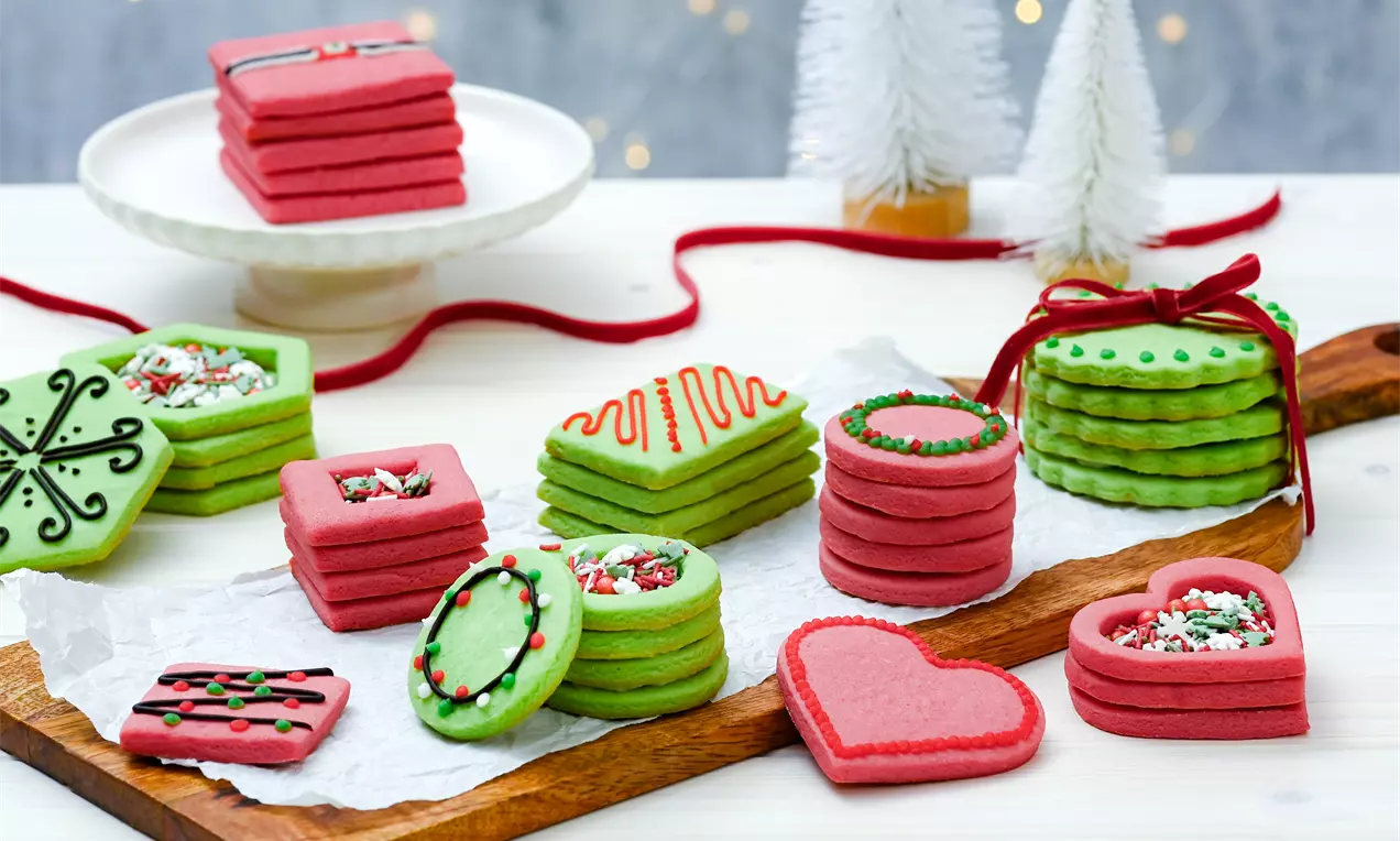 Коледни бисквити в червено и зелено с глазура и декорации, подредени върху бяла хартия върху дървена подложка, със снежни дървета на заден план