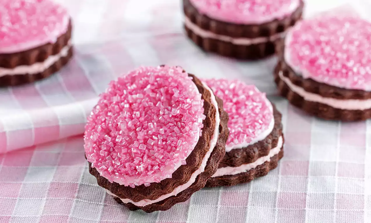 Runde Sandwich-Cookies aus dunklem Teig mit rosa Zuckerdekor und heller Cremefüllung auf einem rosa-weiß karierten Tuch arrangiert