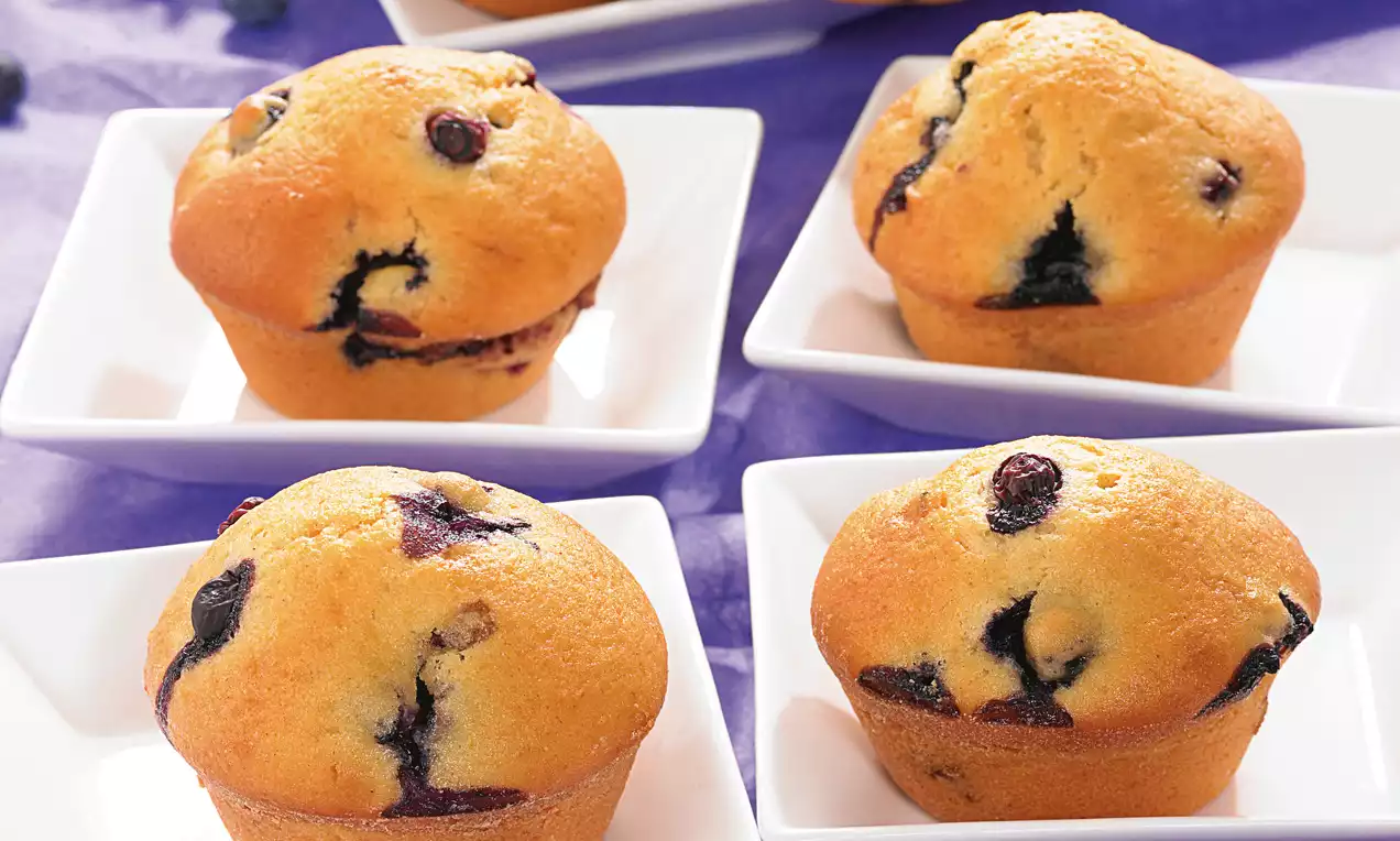 Kullanruskeita, pyöreämuotoisia mustikkamuffineja, joissa syvänsinisiä mustikoita ja kohokuvioinen pinta, aseteltuna valkoisille neliölautasille