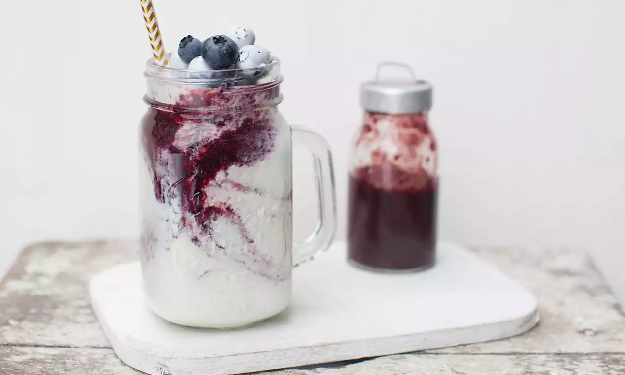 Blueberry platte kaas in een glazen mason jar met paarsrode bessensaus, blauwe bessen en rietje, op een wit bord met confiturepotje ernaast